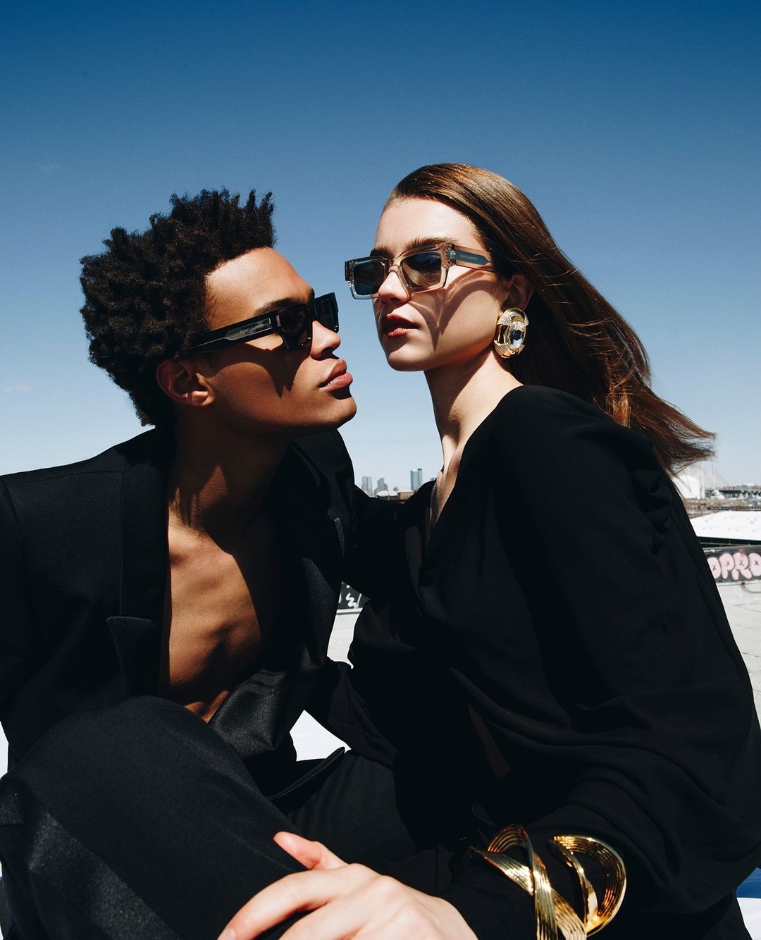 Two stylish models, a man and a woman, pose outdoors in black clothing and sunglasses against a blue sky with city skyline backdrop.