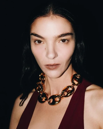 Close-up portrait of a female model wearing a burgundy dress and a chunky gold necklace, set against a dark, high-contrast background.