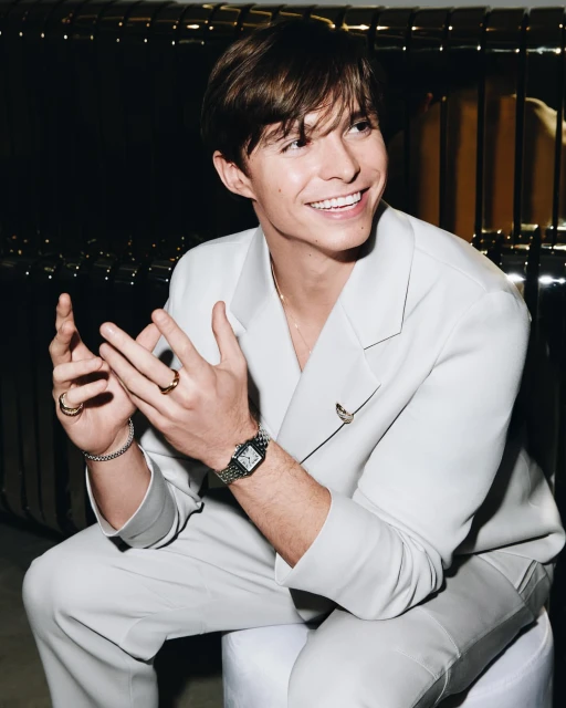A young man in a light suit smiles with hands together, showcasing rings and a watch, set against a dark background. Light is soft and neutral.