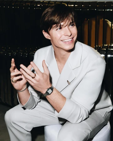 A young man in a light suit smiles with hands together, showcasing rings and a watch, set against a dark background. Light is soft and neutral.