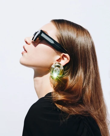 Profile portrait of a woman wearing black sunglasses and a large statement earring against a white background