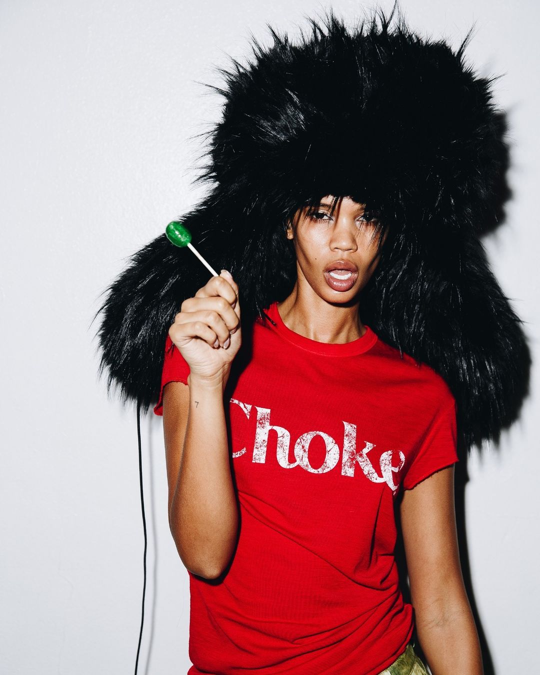 Portrait of an adult woman wearing a black furry hat and red t-shirt, holding a green lollipop in her hand against a plain wall.