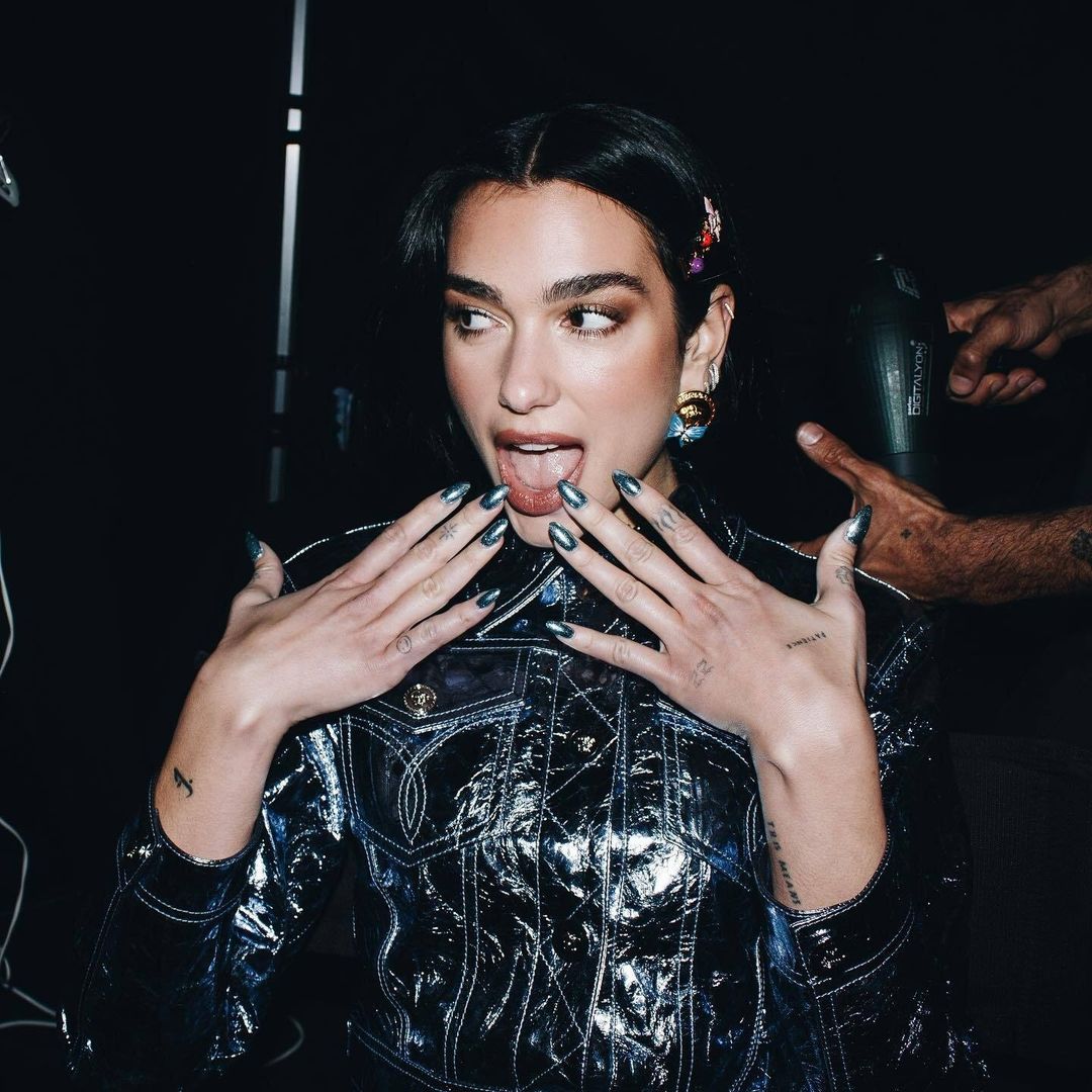 Dua Lipa poses with a playful expression, displaying her silver-blue nails while standing indoors, and wearing a shiny, dark blue jacket.