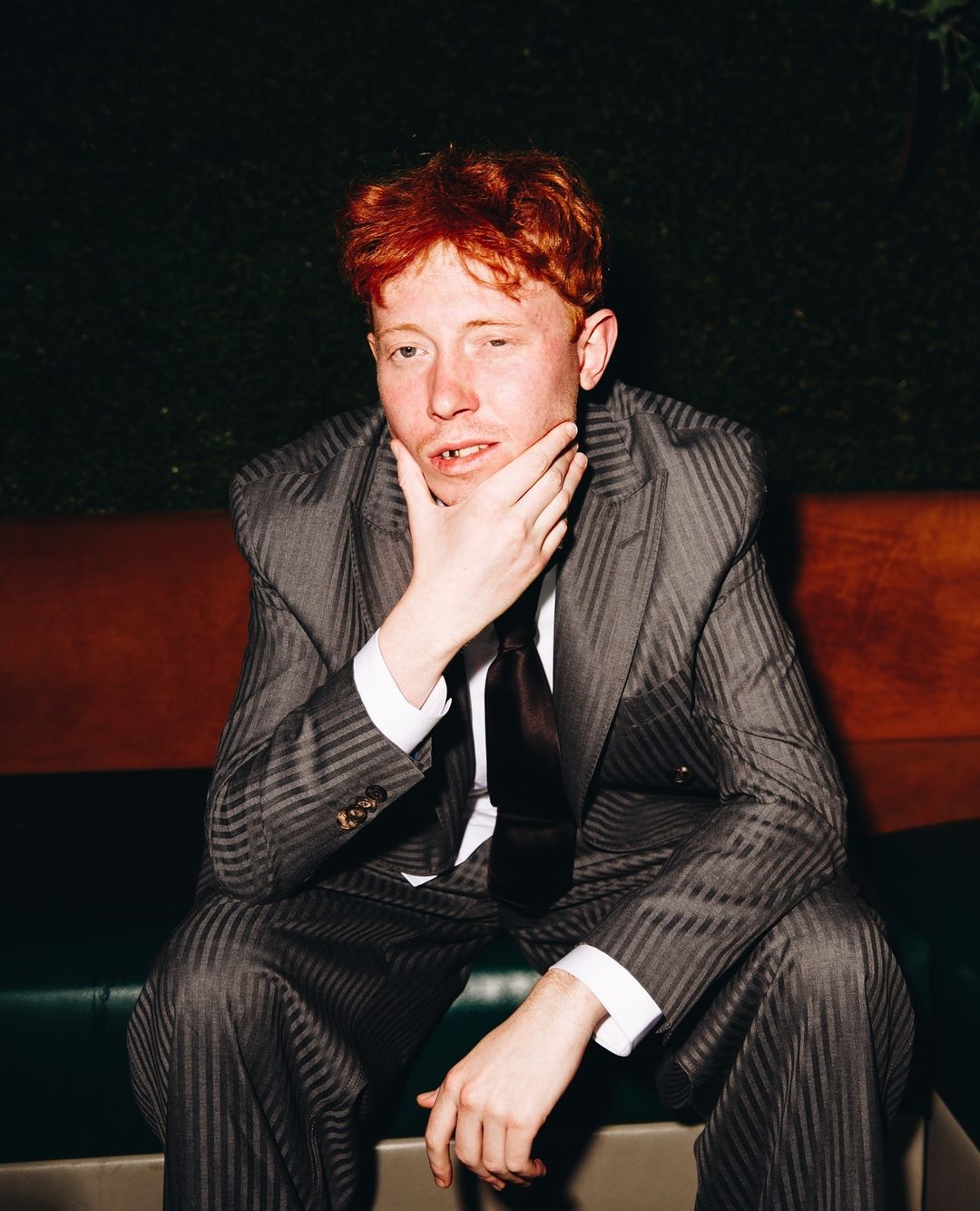 Adult male with reddish hair sits in a patterned suit indoors, hand on his chin, gazing at the camera with a neutral expression.