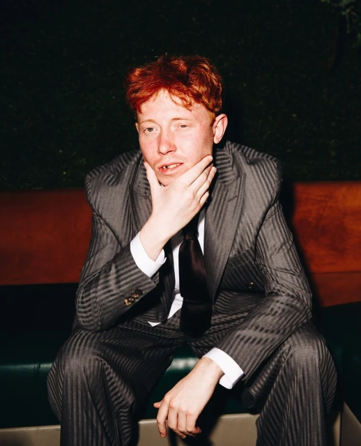 Adult male with reddish hair sits in a patterned suit indoors, hand on his chin, gazing at the camera with a neutral expression.