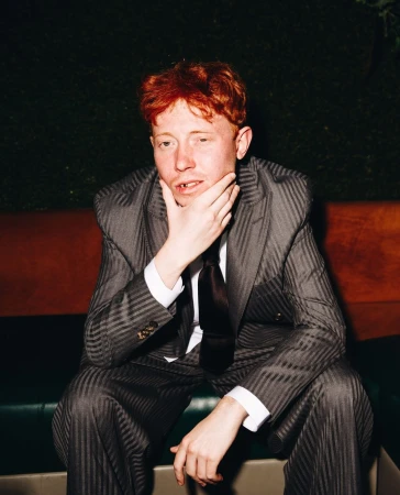Adult male with reddish hair sits in a patterned suit indoors, hand on his chin, gazing at the camera with a neutral expression.