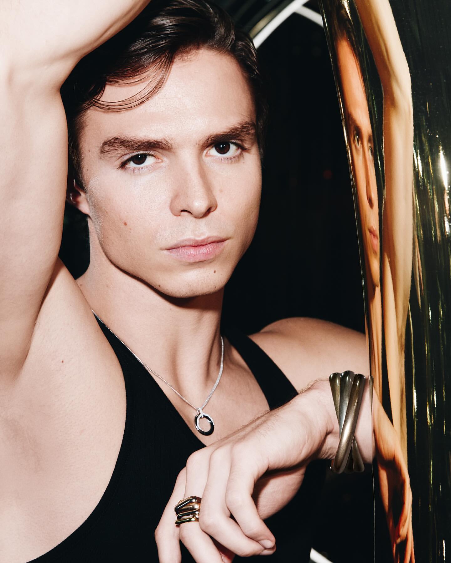 Close-up studio shot of a young adult male model with dark hair, wearing a black tank top, necklace, and jewelry, leaning on a shiny pole.