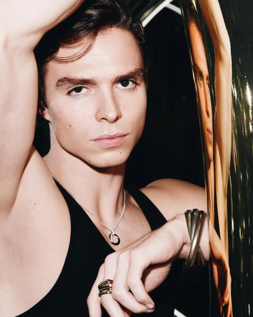 Close-up studio shot of a young adult male model with dark hair, wearing a black tank top, necklace, and jewelry, leaning on a shiny pole.