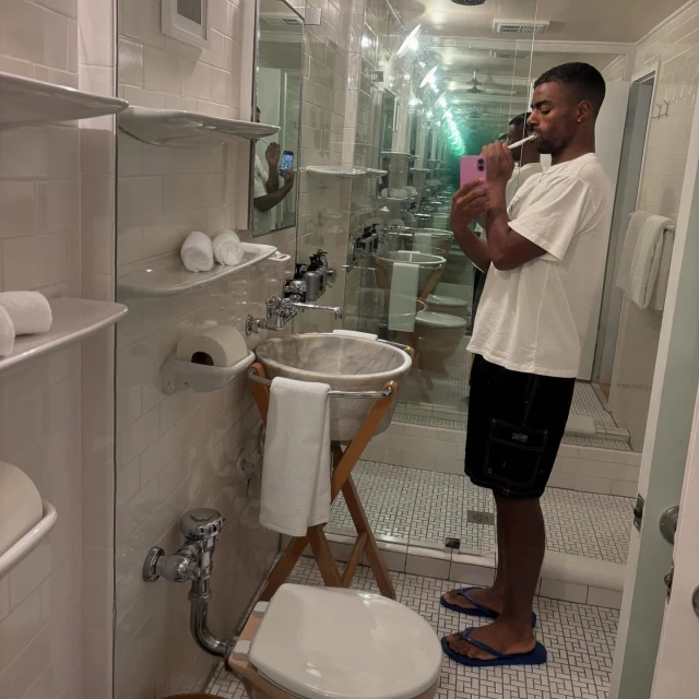 Man in bathroom brushing teeth in front of mirror with infinity effect reflecting toilet, sink, and tiled walls in a brightly lit interior.