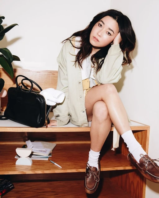 Adult woman with dark hair sitting on a shelf with a bag and plant, wearing a light jacket, white top and socks, and brown loafers.