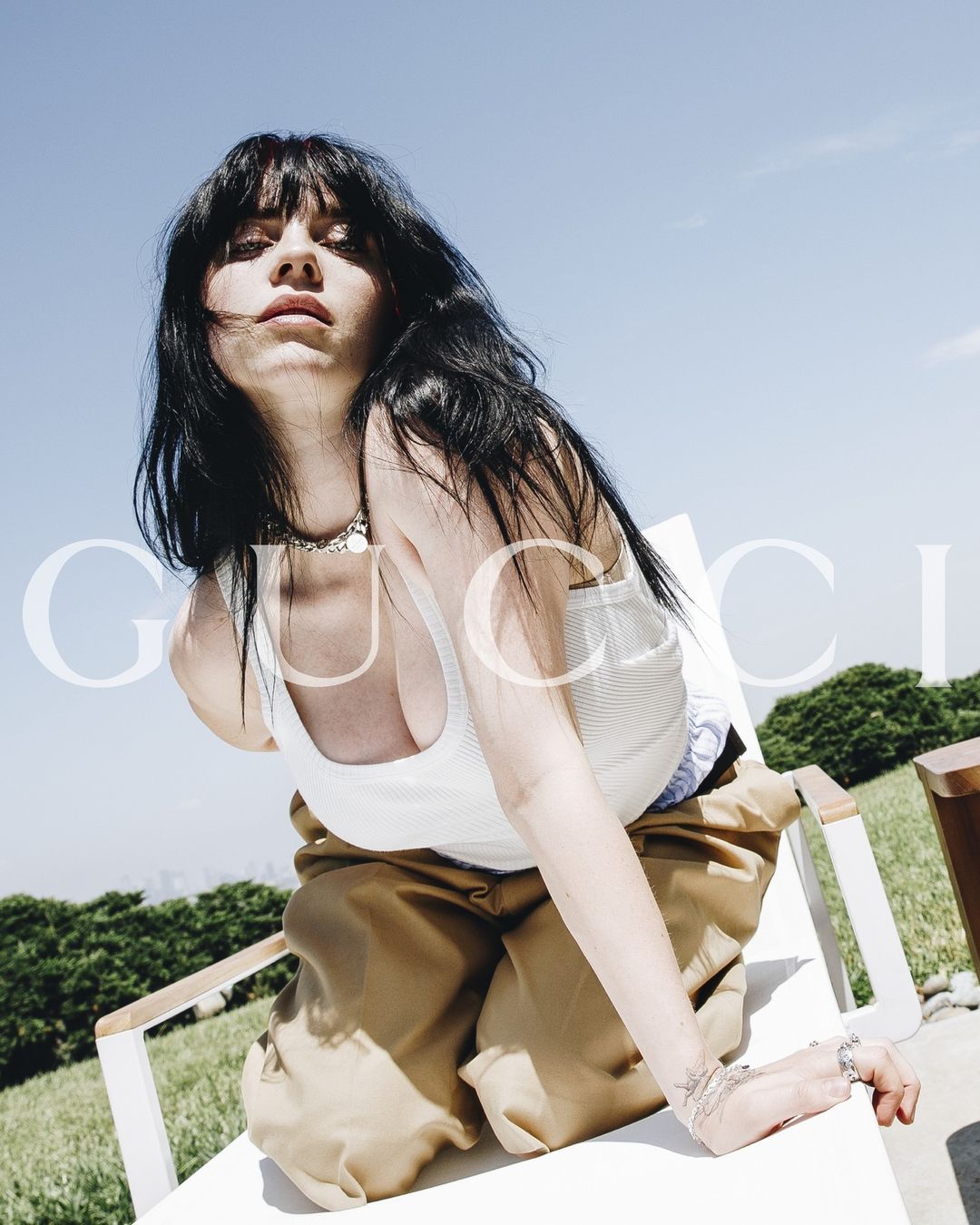 Brunette woman poses outdoors in tank top and loose pants with Gucci logo watermark, shot at eye-level under blue sky.