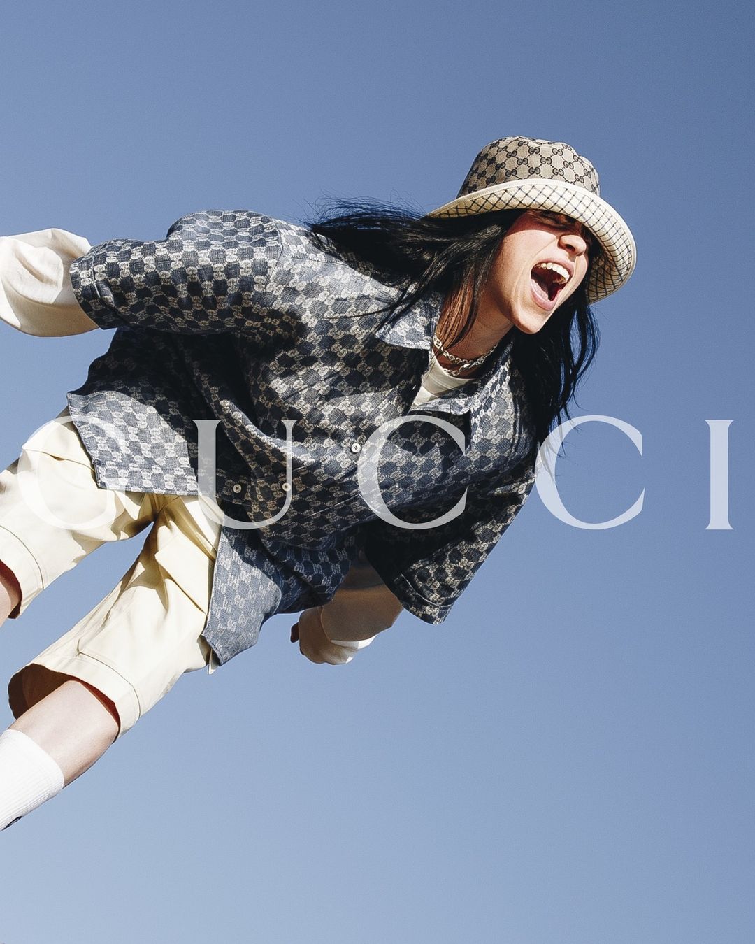 Young woman with open mouth and long dark hair wears patterned shirt and hat in front of light blue sky, Gucci logo overlayed.
