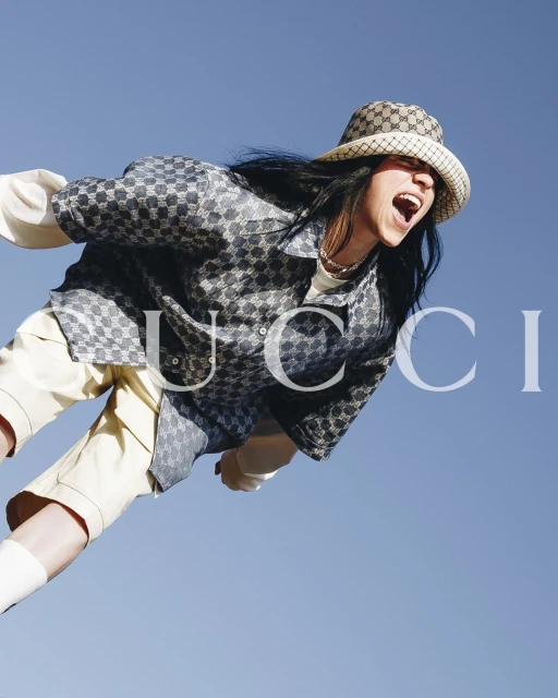 Young woman with open mouth and long dark hair wears patterned shirt and hat in front of light blue sky, Gucci logo overlayed.