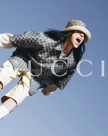 Young woman with open mouth and long dark hair wears patterned shirt and hat in front of light blue sky, Gucci logo overlayed.