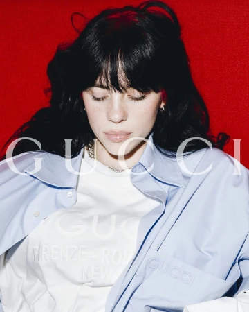 Billie Eilish with dark hair wearing a light blue button-down shirt and white t-shirt against a red backdrop, photographed by Tyrell Hampton