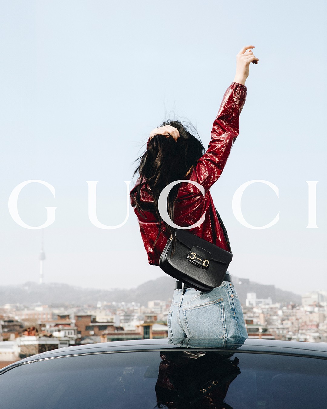 Woman in red jacket and jeans poses on car roof against city. Black purse, blue sky, cityscape, and 'Gucci' brand logo visible.