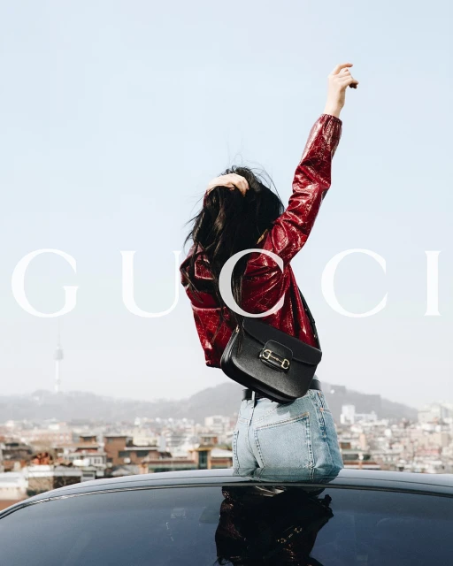 Woman in red jacket and jeans poses on car roof against city. Black purse, blue sky, cityscape, and 'Gucci' brand logo visible.