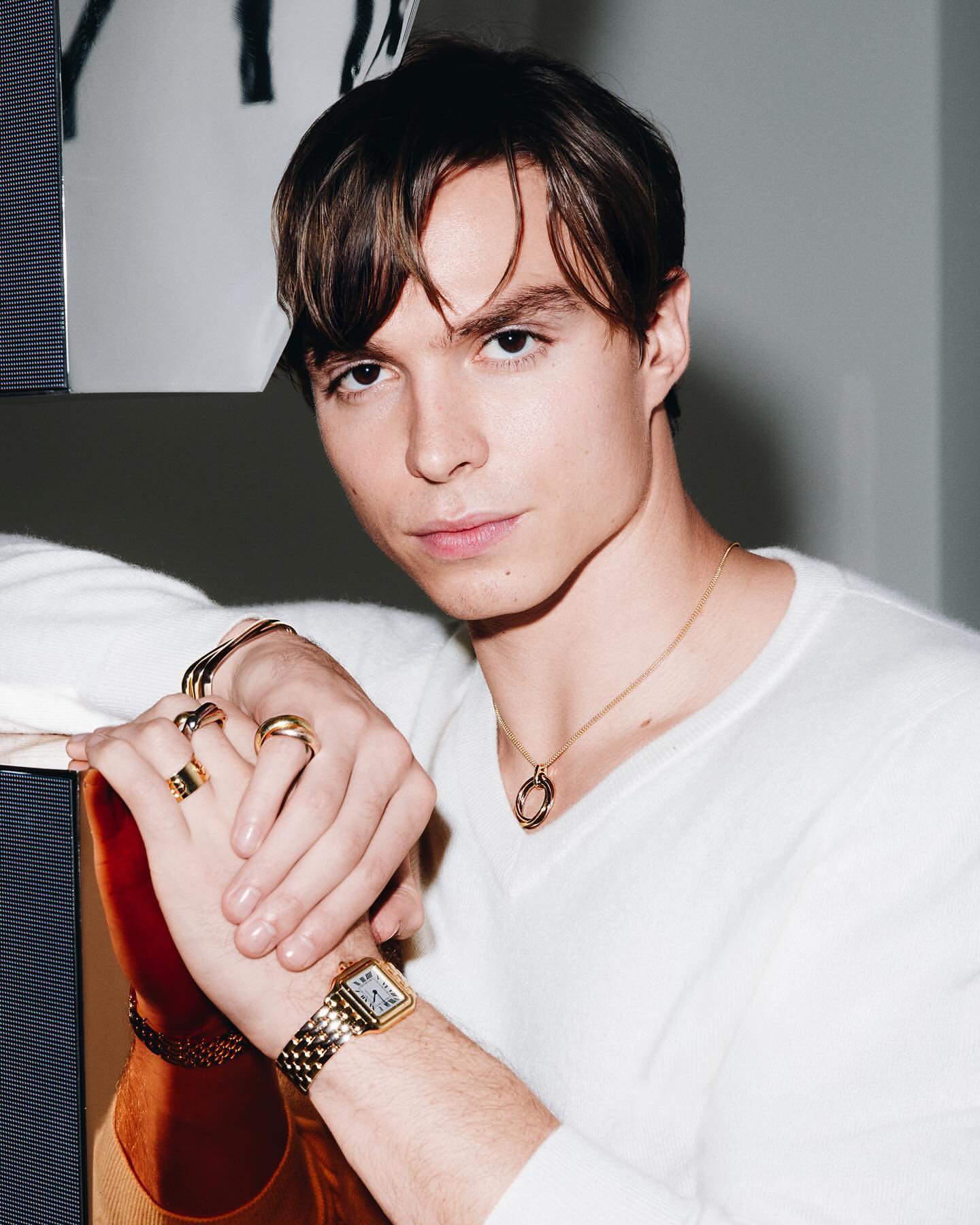 Portrait of a young man with brown hair wearing gold jewelry and a white sweater indoors, with a neutral background, showcasing fashion and style.
