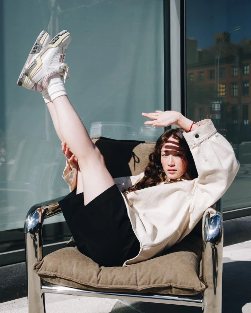 Young woman in a beige jacket and black shorts poses in a modern chair outdoors with her legs up, showing off white sneakers.
