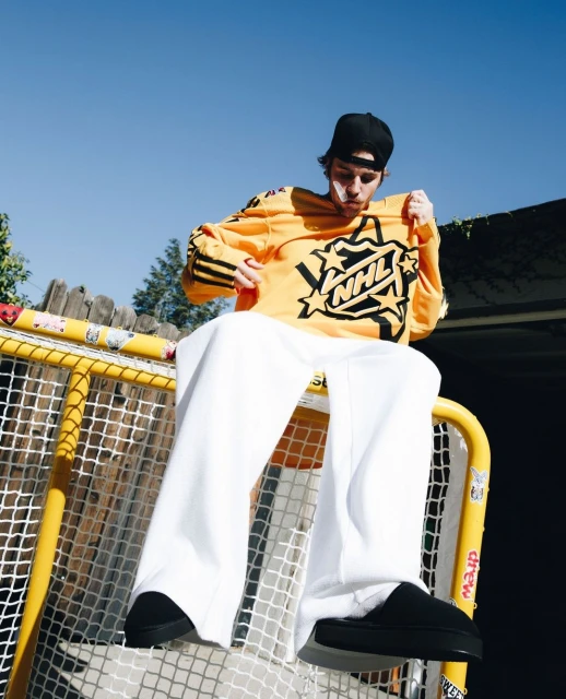 Adult male sits atop yellow hockey goal wearing white pants and black slippers, holding a yellow NHL sweatshirt with black accents.
