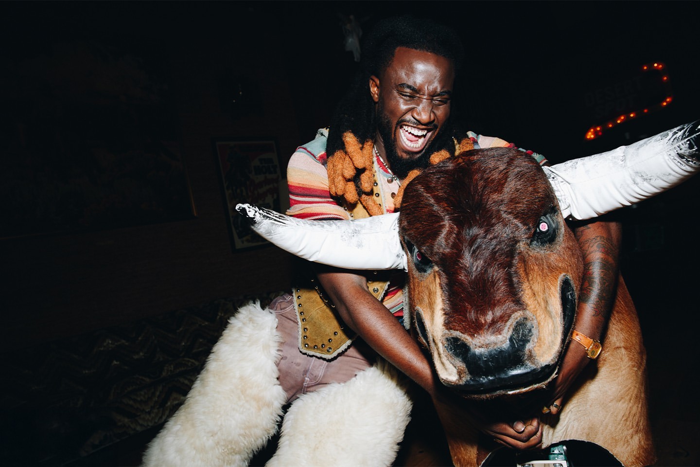 Smiling man rides a mechanical bull indoors at night, wearing a colorful sweater vest and accessorized pants in low lighting.
