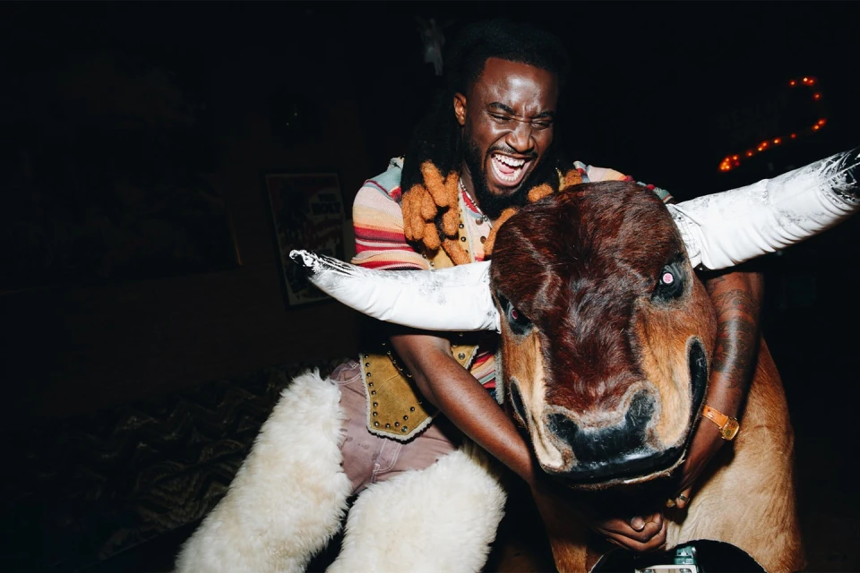 Smiling man rides a mechanical bull indoors at night, wearing a colorful sweater vest and accessorized pants in low lighting.