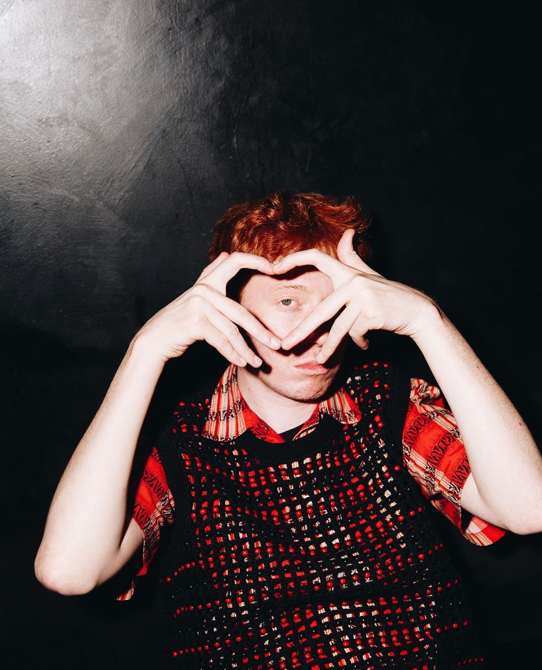 Portrait of a young adult man with red hair in a black and red vest making a heart shape with his hands against a dark background.