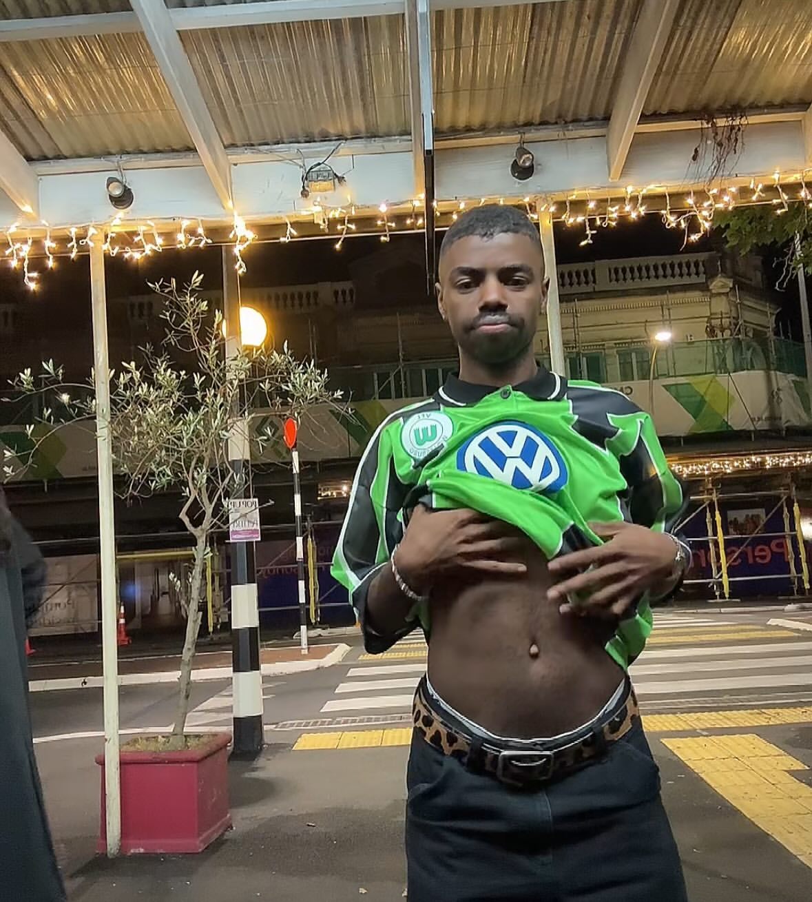A young man stands outside in a green vintage soccer jersey and black pants with a leopard print belt at nighttime.