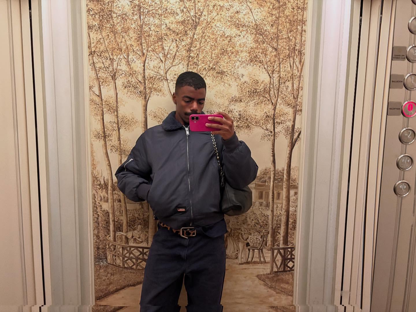 Person in elevator takes selfie with a pink phone wearing a bomber jacket, dark pants, and patterned belt against an illustrated wall.