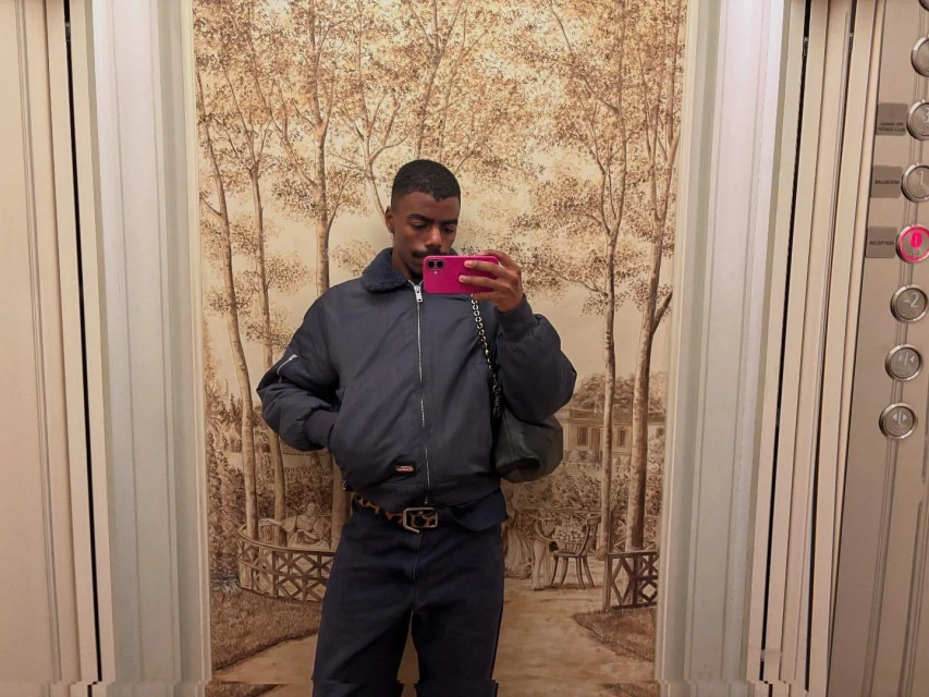 Person in elevator takes selfie with a pink phone wearing a bomber jacket, dark pants, and patterned belt against an illustrated wall.