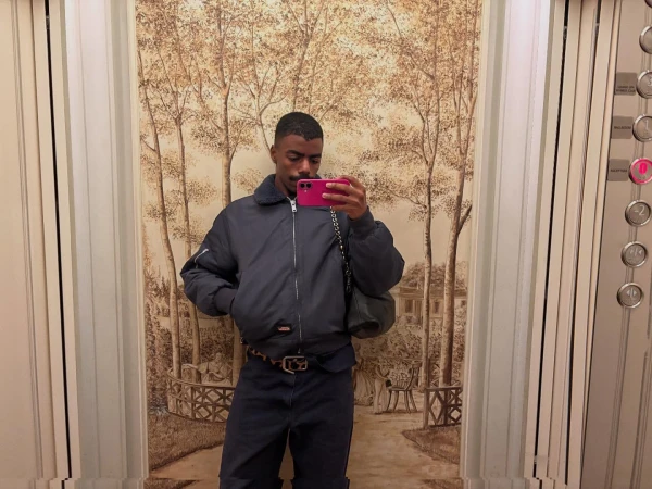 Person in elevator takes selfie with a pink phone wearing a bomber jacket, dark pants, and patterned belt against an illustrated wall.