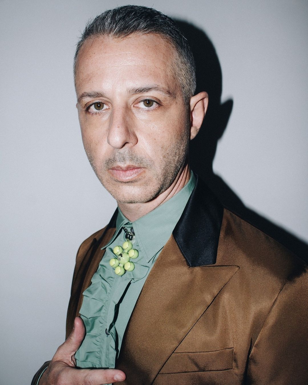 Adult male in a tan suit jacket and light green collared shirt with ruffles, wearing an elaborate brooch, posing against a gray backdrop.
