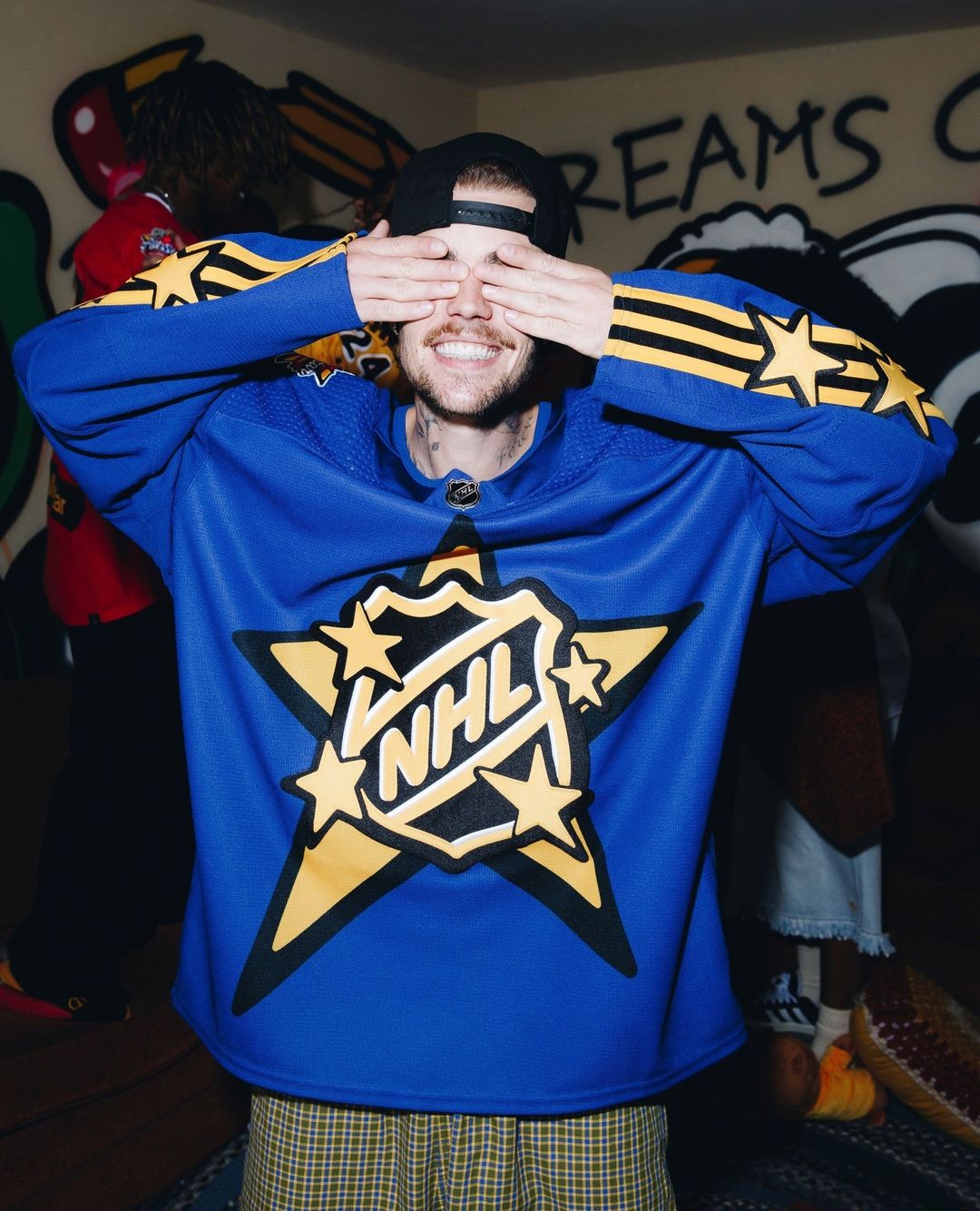 Man in NHL jersey covers eyes with hands, smiling indoors against graffiti wall. Visible blue jersey, star logo, baseball cap, fair skin.