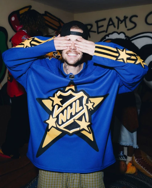 Man in NHL jersey covers eyes with hands, smiling indoors against graffiti wall. Visible blue jersey, star logo, baseball cap, fair skin.