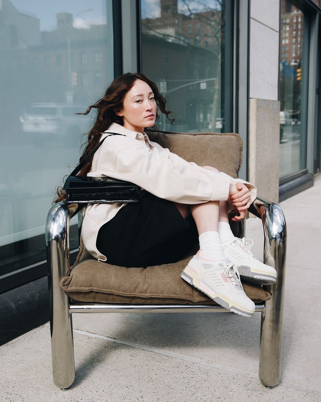 Woman with long dark hair seated in a chrome armchair wearing a light jacket, black shorts, and sneakers against a building exterior.