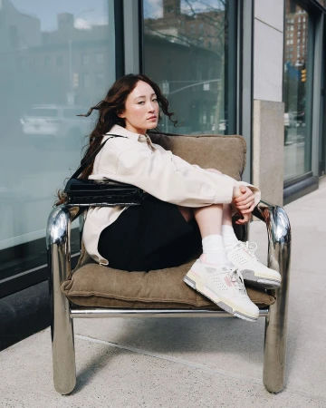 Woman with long dark hair seated in a chrome armchair wearing a light jacket, black shorts, and sneakers against a building exterior.