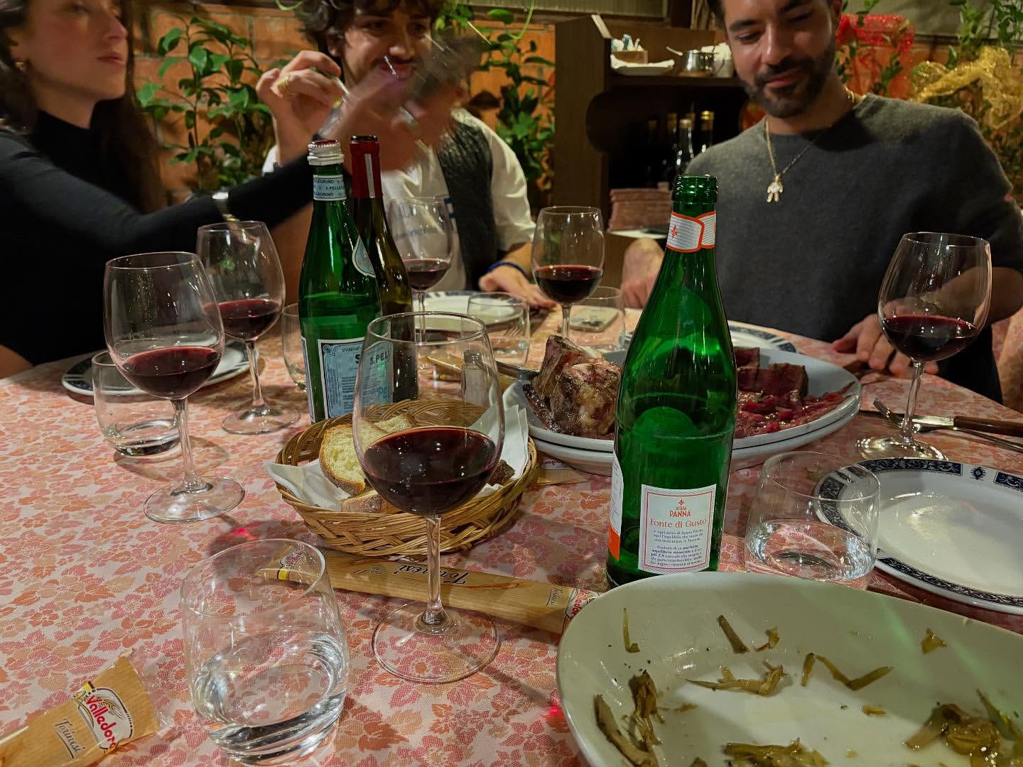 Indoor dinner party with four people gathered around a table laden with wine glasses, bottles of water, bread, and food platters, in warm lighting.