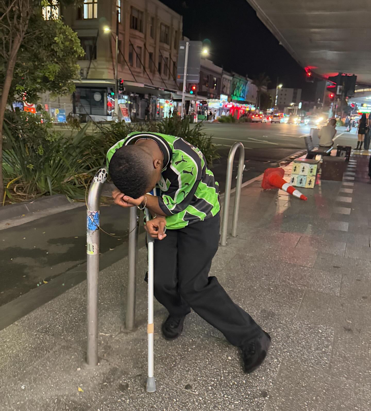Man in green soccer jersey leans on metal railing with white cane on city sidewalk at night. Street lights and buildings in background.