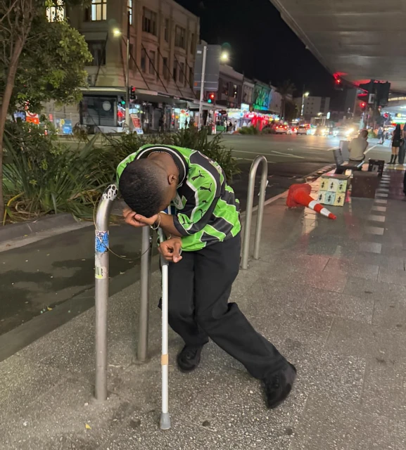 Man in green soccer jersey leans on metal railing with white cane on city sidewalk at night. Street lights and buildings in background.