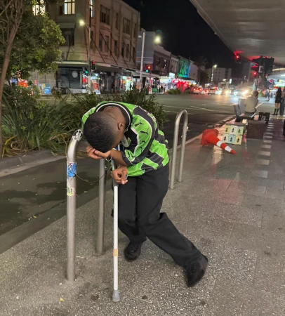 Man in green soccer jersey leans on metal railing with white cane on city sidewalk at night. Street lights and buildings in background.