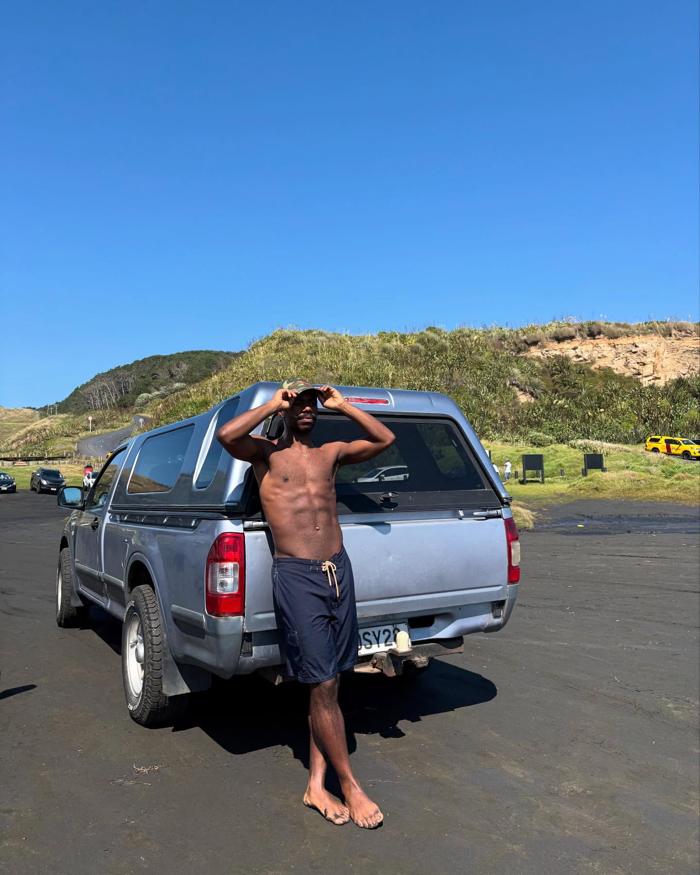Shirtless adult man posing next to a silver pickup truck on a black sand beach with dunes and a clear blue sky in the background.