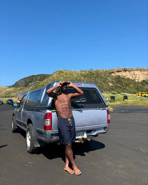 Shirtless adult man posing next to a silver pickup truck on a black sand beach with dunes and a clear blue sky in the background.