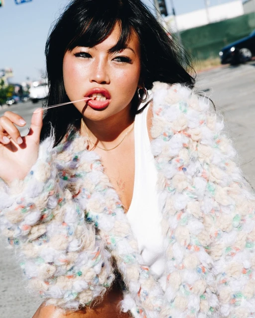 Young Asian woman poses in street with fuzzy pastel jacket, white top, chewing gum, hoop earrings, traffic lights, and dark car.