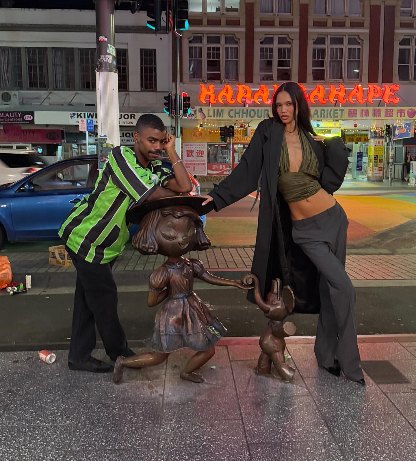 Two young adults, a man and a woman, pose next to bronze child sculptures on a city sidewalk at night, with neon signage in the backdrop.