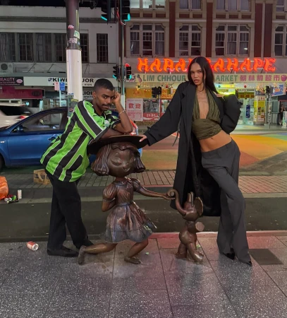 Two young adults, a man and a woman, pose next to bronze child sculptures on a city sidewalk at night, with neon signage in the backdrop.