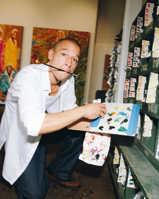 Artist kneels with palette and paintbrush in an art studio, wearing a white shirt and blue jeans, with abstract art and art supplies visible.