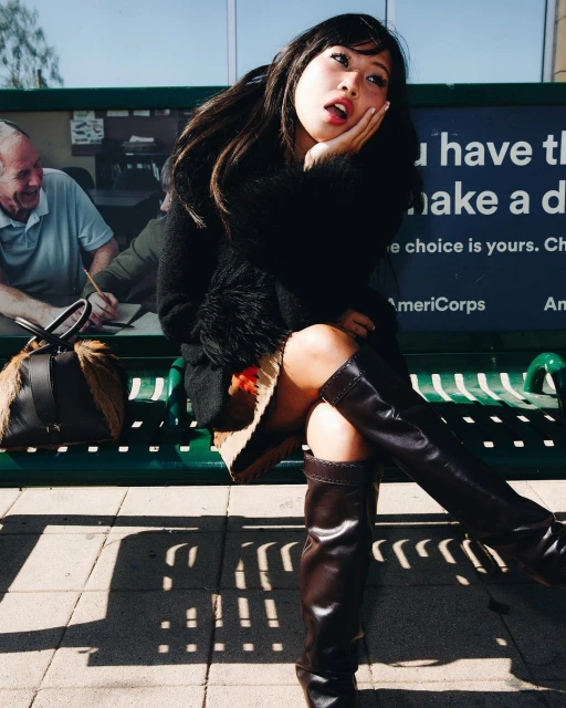 Young Asian woman sits on a bench outdoors in thigh-high leather boots, wearing a black jacket and skirt, with a bag beside her.