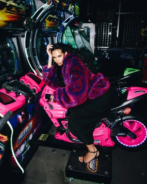 A woman sits on a pink motorcycle arcade game, wearing a colorful faux fur coat, in a dimly lit entertainment space with other games visible.