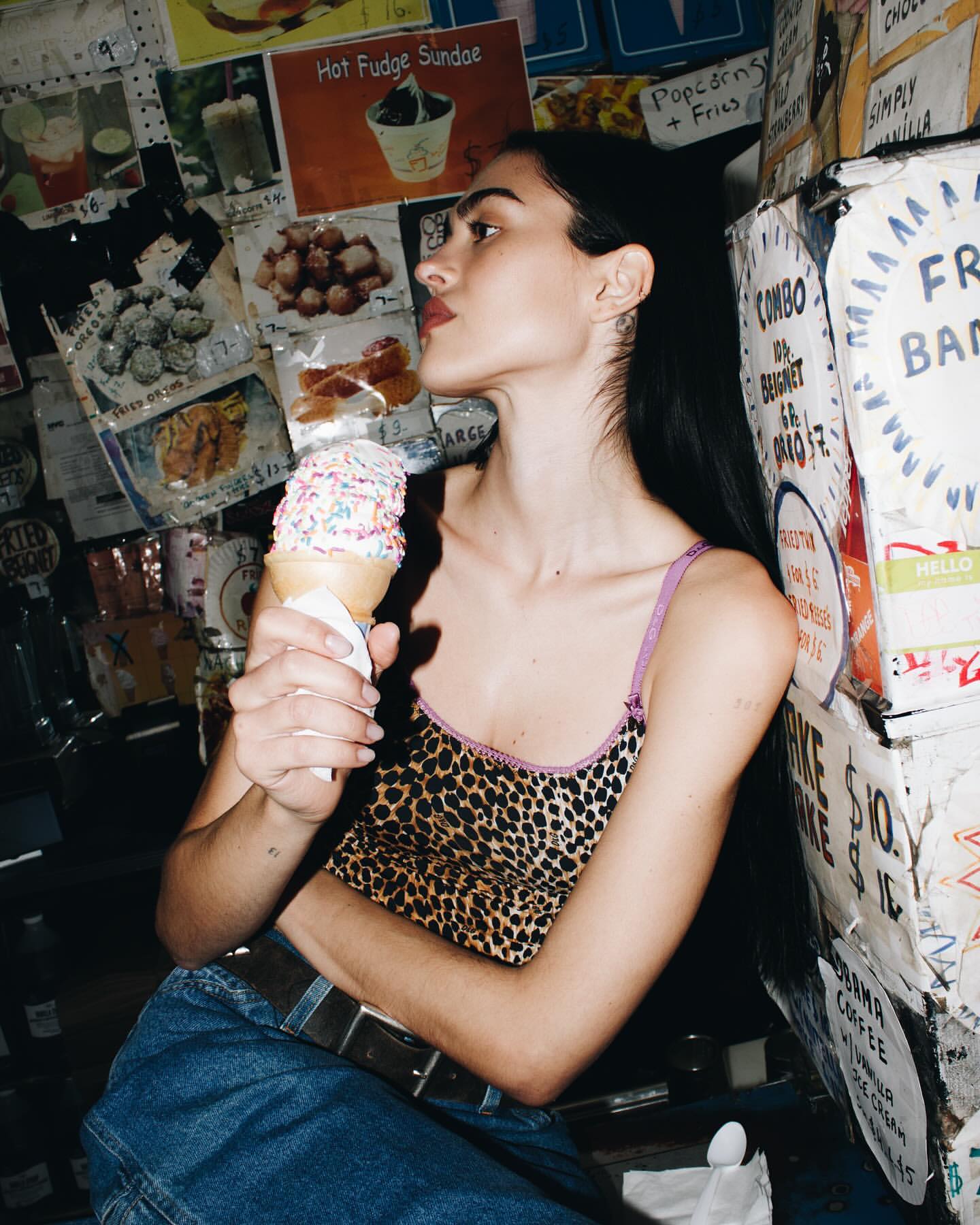 A young woman with long dark hair, wearing a leopard-print top and jeans, eats a sprinkle-covered ice cream cone against a wall of posters.