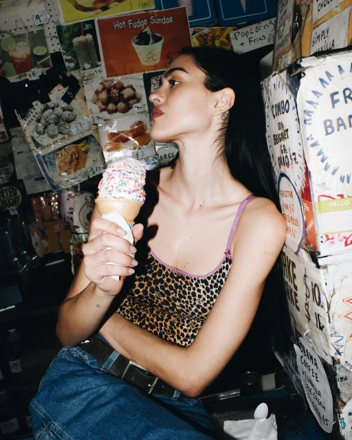 A young woman with long dark hair, wearing a leopard-print top and jeans, eats a sprinkle-covered ice cream cone against a wall of posters.
